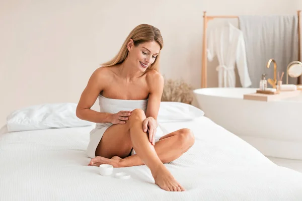 Happy middle aged lady applying moisturizer cream on legs, caring for body and moisturizing skin after shaving, sitting on bed, free space. Aftershave skincare routine concept