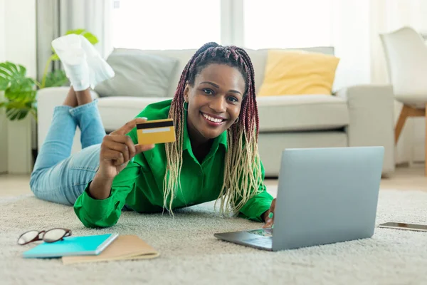 Happy african american lady shopping via laptop and credit card, buying new clothes and making online payment, lying on floor carpet at home. Internet banking and ecommerce concept