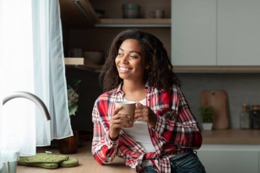 Neşeli bin yıllık Afro-Amerikan kadın bardakla içkinin, rahatlığın ve sükunetin tadını çıkarıyor, minimalist mutfak içlerine, bedava alana bakıyor. Kahve molası, evde dinlenme ve rahatlama, covid-19