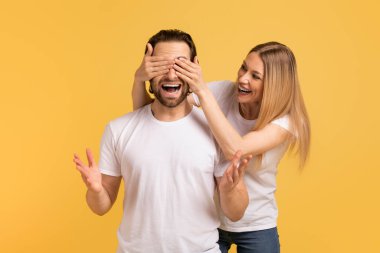Beyaz tişörtlü, gülümseyen beyaz kadın sarı arka planda izole edilmiş ağzı açık, şok olmuş adama gözlerini kapar. Duygular, doğum günü sürprizi, haberler, harika reklam ve teklif, aşk, ilişki