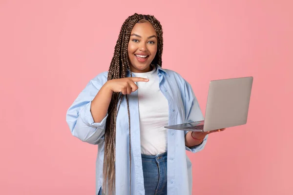 Cheerful African American Woman Using Laptop Pointing Finger At Computer Working Online And Browsing Internet Posing Standing Over Pink Background. Studio Shot