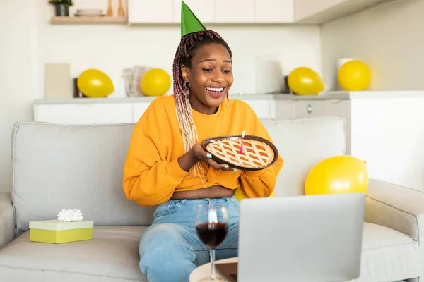 Excited black lady holding birthday pie with candle, video calling via laptop, having remote celebration at home, sitting on sofa. Online b-day party concept