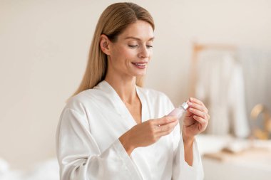 Serum kullanan mutlu orta yaşlı kadın, modern banyo içinde cilt bakım ev. Yüz bakımı, güzellik tedavisi rutin konsept.