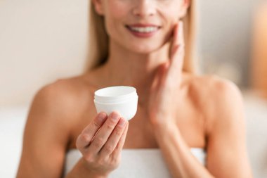Skin care concept. Unrecognizable middle aged woman holding jar with moisturizing cream and applying nourishing lotion on her face, selective focus, cropped clipart