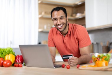 Masadaki organik sebzeli neşeli siyah erkek minimalist mutfak içinde dizüstü bilgisayar ve kredi kartı kullanıyor. Çevrimiçi alışveriş için araç, covid-19 salgını sırasında evde uzaktan satış