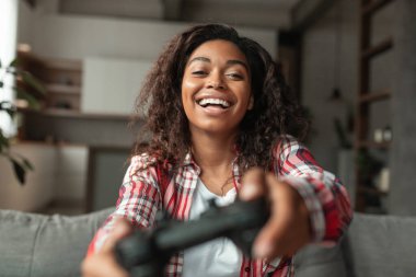 Mutlu bin yıllık Afro-Amerikan kadın kanepede oturup joystickle online oyun oynayarak oturma odasında boş vakitlerin tadını çıkarıyor. Rahat evinizde dinlenin, teknolojiyle hobileriniz ve eğlenceniz