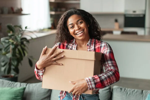 Satisfied pretty millennial african american lady hugs cardboard box with purchases in living room interior. Shopaholic enjoy sale, delivery and online shopping, new normal and emotions, ad and offer