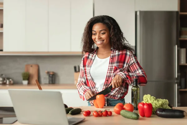 Cheerful millennial african american lady cuts organic vegetables and looks at laptop, enjoys cooking food in minimalist kitchen interior. Technology, blog, new salad recipe, veganism and health care