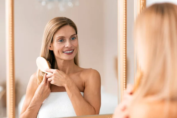 Daily beauty routine. Happy middle aged woman combing her hair with brush while standing near mirror. Attractive mature lady looking to her reflection and smiling