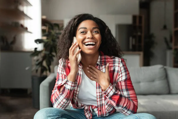 Laughing millennial black lady with open mouth sitting on sofa, talking on phone in living room interior. Call and communication at home, gossip and good news at leisure time, emotions, ad and offer