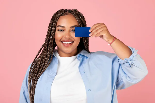 Cheerful Black Female Holding Credit Card Covering Eye Having Fun Posing Smiling To Camera Standing On Pink Background. Great Bank Offer Concept. Studio Shot