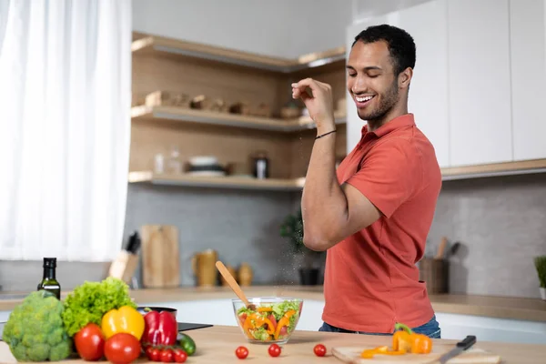 Happy funny millennial african american guy salts salad, prepare food at table with organic vegetables in kitchen interior. Household chores, cooking for family, health care and veganism at home