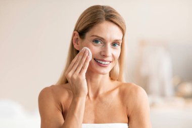 Cilt rutini kavramı. Mutlu orta yaşlı kadın pamukla cildi temizliyor, makyajı siliyor, beyaz havluya sarılıp kameraya gülümsüyor.