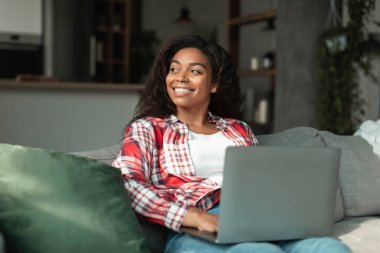 Bin yıllık gülümseyen, dizüstü bilgisayarlı Afro-Amerikan kadın boş alana bakıyor, oturma odasının iç tarafındaki kanepede oturuyor. Harika bir reklam ve teklif, satış, insan duyguları, iş ve iş vadesi covid-19.