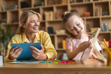 Zeki küçük kız sınıfta öğretmenle egzersiz yapıyor, alfabe öğreniyor ve gemide kelimeler yazıyor, profesyonel öğretmen öğrenciyi izliyor ve notlar alıyor.