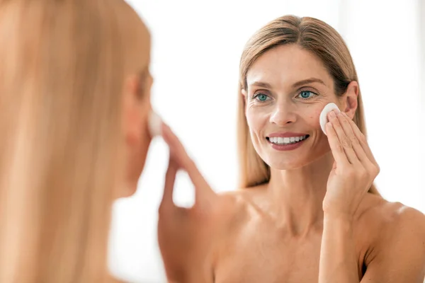 Morning beauty routine. Smiling middle aged woman cleansing face with cotton pad and looking in mirror, mature lady doing anti-aging skincare treatments after shower