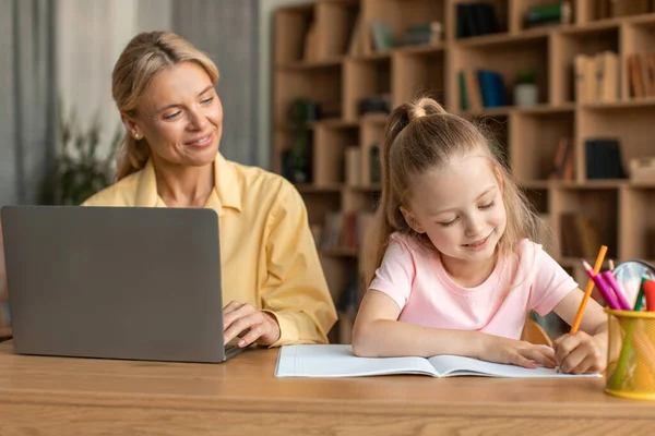 Neşeli anne dizüstü bilgisayar kullanıyor, uzaktan çalışıyor ve kızıyla vakit geçiriyor, küçük tatlı kız çiziyor ya da ödevlerini yapıyor, birlikte masada oturuyor.
