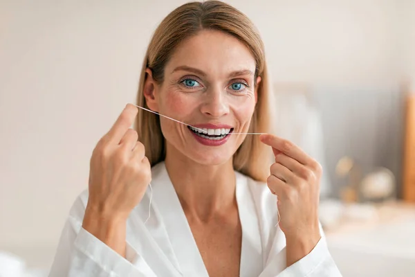 Oral health concept. Middle aged woman using dental floss, making daily hygiene, wearing bathrobe, posing in bathroom interior, looking and smiling at camera