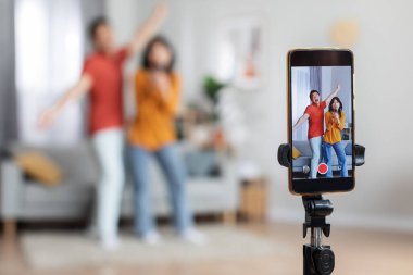 Mutlu Asyalı karı koca sosyal medya veya kişisel aile videoları için video kaydediyor, kameranın önünde el ele tutuşup dans ediyor, tripodlardaki akıllı telefon setine odaklanıyor, ev içi