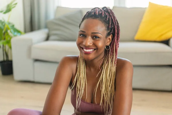 Portrait of black woman in sportswear posing and smiling to camera, sitting on yoga mat in living room interior, taking break from domestic workout