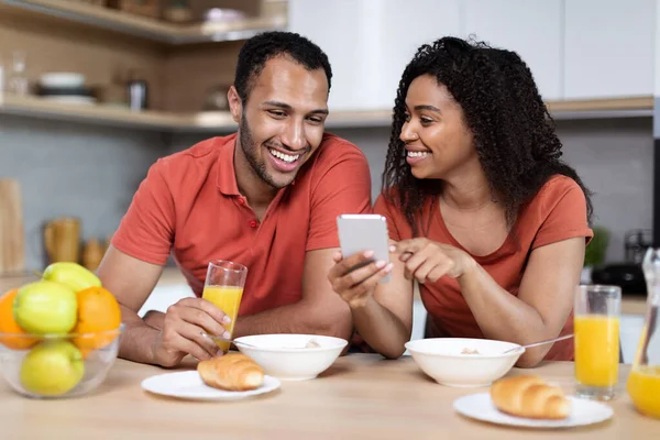 微笑的千年黑人夫妇喜欢早上一起在简约的厨房里看智能手机吃早餐 在家里远程呼叫视频的新应用和小玩艺儿 广告和提供 聊天和博客 — 图库照片