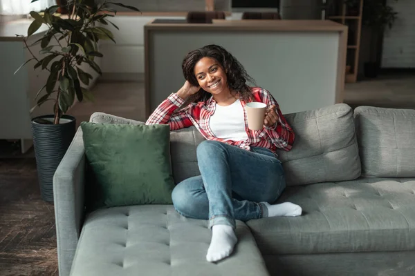 Cheerful millennial african american lady with cup of favorite drink, enjoy free time and comfort, sit on sofa in living room interior. Human emotions, rest and relax in cozy home, coffee break alone