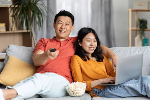 Relaxed chinese lovers resting together at weekend at home, happy asian middle aged man watching TV, hugging young woman using laptop, sitting on couch, eating popcorn, copy space