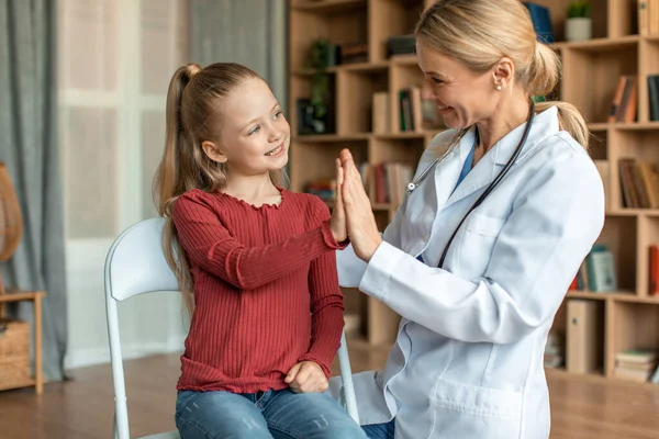 Little girl at appointment with friendly female doctor, healthy happy cute kid giving pediatrician high five and smiling, home interior