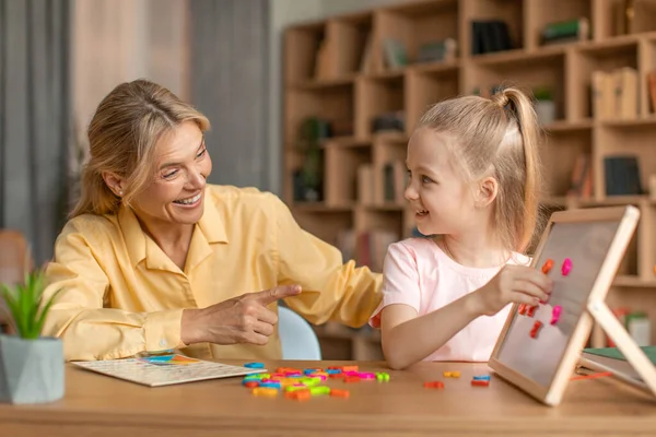 Excited woman speech therapist teaching adorable little girl alphabet, making words from plastic letters on whiteboard. Speech-language therapy for children concept