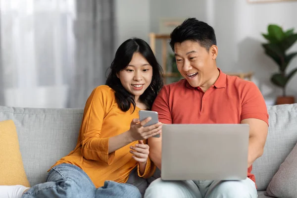 Positive loving japanese middle aged man and young woman sitting on couch at home, using gadgets cell phone and computer, having conversation, copy space. Gadget addiction and relationships