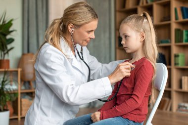 Kadın doktor, evdeki sağlık kontrolü sırasında çocuğun ciğerlerini kontrol ediyor. Dost canlısı bayan çocuk doktoru steteskop kullanarak genç bir hastanın nefes alışını ve kalp atışlarını inceliyor.