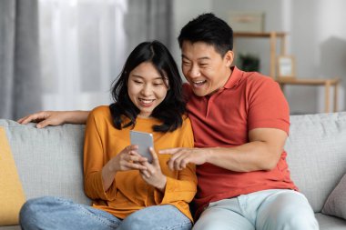 Mutlu Asyalı aşıklar evde birlikte vakit geçiriyorlar, pozitif orta yaşlı erkek ve genç kadın oturma odasında kanepede dinleniyorlar, sarılıyorlar ve akıllı telefon kullanıyorlar, mobil oyun oynuyorlar, uzay kopyaları yapıyorlar.