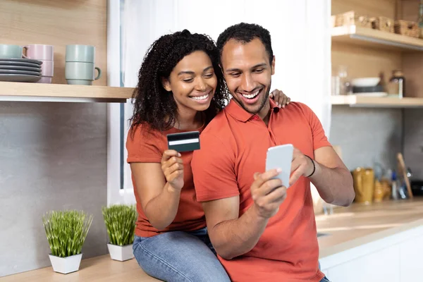 Cheerful millennial black lady hugging guy with credit card and smartphone enjoy online shopping in kitchen interior, copy space. Shopaholic couple have fun at sale remotely with modern device and app
