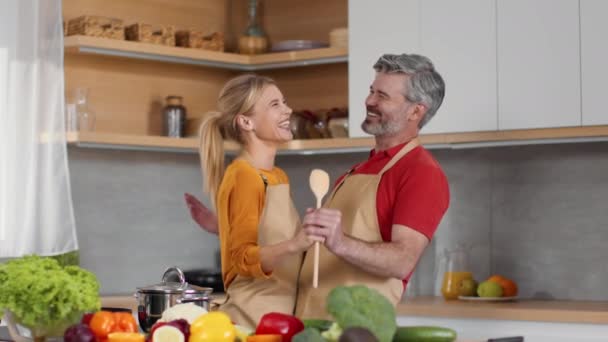 面白い配偶者料理 幸せな笑い中年の既婚男性と女性一緒に踊り キッチンで夕食を調理しながら大きな木製のスプーンに歌います スローモーション — ストック動画