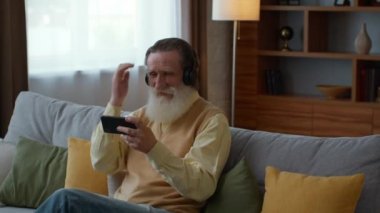 Kıdemli oyuncu. Kulaklıklı duygusal yaşlı sakallı büyükbaba akıllı telefondan video oyunları oynuyor, evde sanal eğlencenin tadını çıkarıyor, çekim takip ediyor, yavaş çekim, boş alan