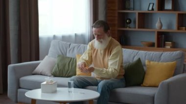 Sağlık için vitaminler. Yaşlı sakallı adam evde ilaç alıyor. Sağlığını takviyelerle, izleme çekimleriyle, yavaş çekimle, boş alanla önemsiyor.