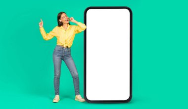 Gülümseyen Avrupalı kulaklıklı genç bir kadın, yeşil arka planda, stüdyoda izole edilmiş, büyük akıllı telefonun yanında eğleniyor, şarkı söylüyor, müziğin tadını çıkarıyor. Reklam ve teklif, web sitesi ve rahatlama için yeni uygulama