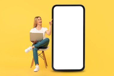 Mutlu Avrupalı genç kadın dizüstü bilgisayarla sandalyede oturuyor, boş ekranlı akıllı telefonu işaret ediyor, sarı arka planda, stüdyoda izole edilmiş. Çevrimiçi iş, reklam ve teklif, İnternet sitesi ve eğitim uygulaması