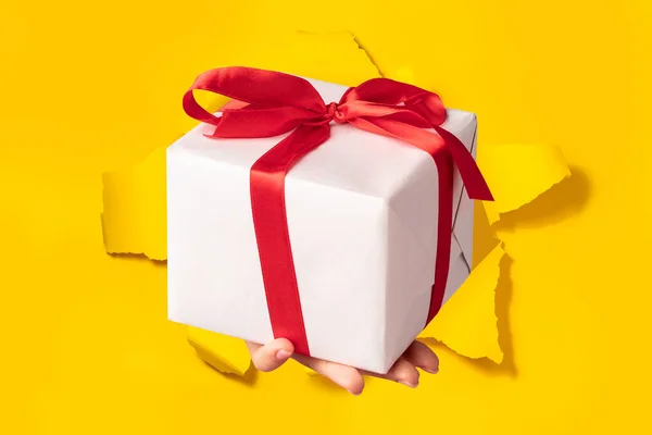 手递礼物盒穿过撕破的黄纸背景 用红色丝带弓以白色包裹的形式出现 特殊报价或销售概念 — 图库照片