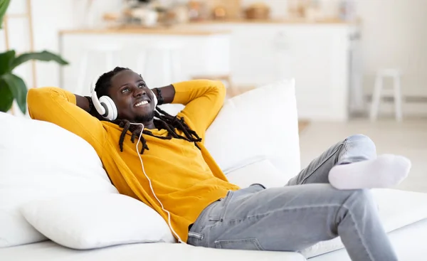 休息だ リビングでソファでリラックスしてヘッドフォンを身に着けている若い黒人男性を笑顔 幸せな千年紀のアフリカ系アメリカ人男性お気に入りの音楽を聞いて 頭の後ろに手でソファにもたれて — ストック写真