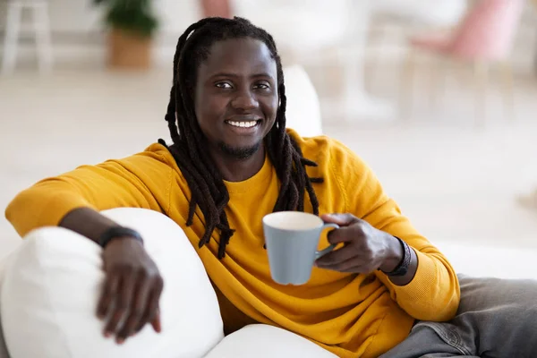 国内パスタイム コーヒーを片手にカップでリラックスしてカメラで微笑む幸せな若い黒人男性 ドレッドロックヘアスタイルで陽気なアフリカ系アメリカ人男性居心地の良いリビングルームで休憩 — ストック写真