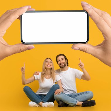 Mutlu Avrupalı kadın ve erkek, sarı arka plan, stüdyo, kolaj gibi izole edilmiş ellerinde boş ekranla büyük akıllı telefonu işaret ediyor. Yeni uygulama, harika teklif, reklam ve büyük indirim.