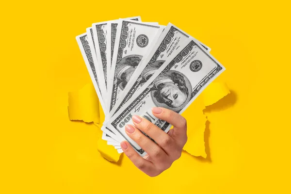 Female hand holding fan of cash money in dollar banknotes, isolated through torn yellow background, closeup. Big profit and financial success