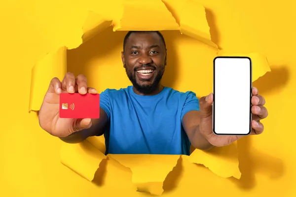 移动购物应用程序 兴奋的黑人男子拿着空白屏幕的信用卡和手机 从黄色背景的洞口往外窥视 带有复制空间的小工具 用于模拟 数字支付 — 图库照片