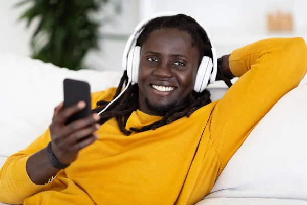 Przystojny Młody Czarny Gość Smartfonem Słuchawkami Relaks Kanapach Domu Uśmiechnięty — Zdjęcie stockowe