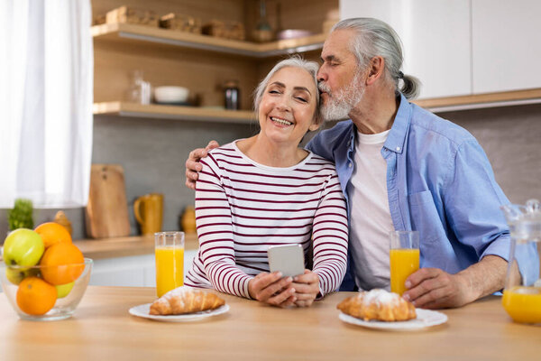 Portrait Of Romantic Happy Senior Couple Resting With Smartphone And Having Breakfast In Kitchen, Cheerful Elderly Spouses Eating Snacks And Using Mobile Phone Together At Home, Husband Kissing Wife