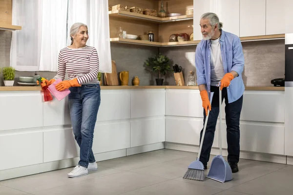 キッチンで一緒に掃除をする幸せ先輩配偶者の肖像 笑顔の高齢夫と妻が家事をする 夫が床を潮吹きしながらテーブルのためにほこりを拭く女 — ストック写真