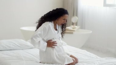 Hamilelik ve sağlık sorunları. Akut karın ağrısı çeken genç, duygusal hamile bir kadın karnına dokunuyor ve çığlık atıyor, evde yatakta oturuyor, ağır çekim, boş alan.