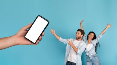 Heyecanlı genç Avrupalı ve Arap aile fertleri ellerinde boş ekranla akıllı telefonu işaret ediyor, mavi arka planda, panoramada, stüdyoda izole edilmiş. Yeni uygulama, teknoloji, harika bir teklif reklamı ve büyük bir indirim.