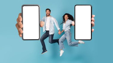 Gülümseyen genç Avrupalı ve Arap aile sıçrayışları, başarının sevinci, boş ekranlı büyük akıllı telefonlar, mavi arka planda izole edilmiş stüdyolar. Zafer online, yeni uygulama, harika teklif, reklam ve satış